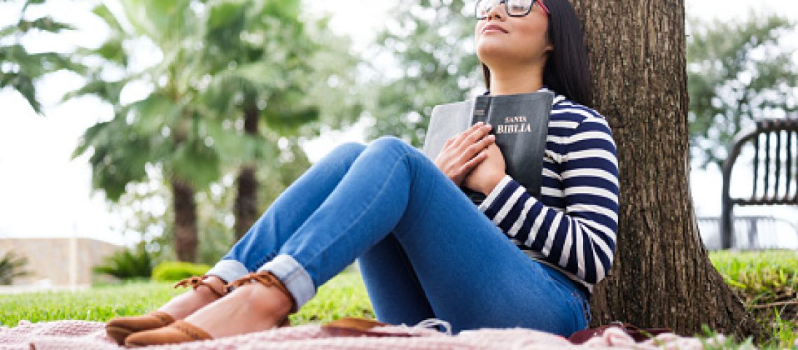 Pretty young woman sitting next to a tree and holding a bible close to her chest.